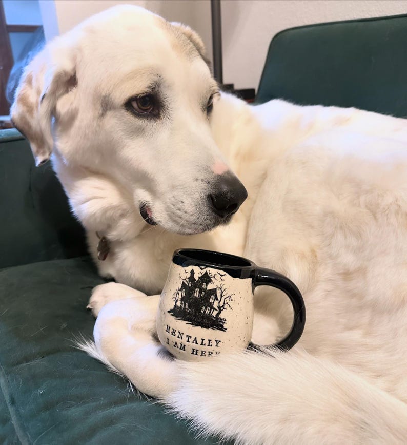 Cassio, a 100 lb. akbash dog, curled up with a mug. The mug is a white speckled clay mug that reads “MENTALLY I AM HERE” in black letters with a black image of a haunted house. The interior, handle, and rim are glazed glossy black.