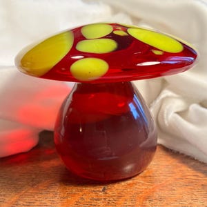 Pode incluir: Uma figura de cogumelo de vidro com um caule vermelho profundo e um chapéu largo e arredondado. O chapéu é de vidro vermelho translúcido decorado com manchas circulares amarelo-brilhantes, lisas e de vários tamanhos na superfície.