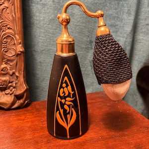 May include: An antique black perfume bottle with a gold-colored atomizer and a black woven bulb. The bottle has an orange floral design. The bottle is on a wooden surface.