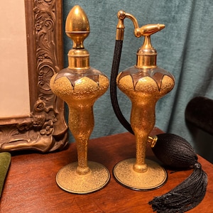 May include: Two antique perfume bottles with gold-colored metal bases and tops. The glass bottles have a decorative gold pattern. One bottle has a gold stopper, and the other has a black atomizer with a tassel.
