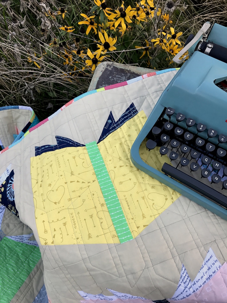 Pu&ograve; includere: Un quilt giallo e verde con un motivo a cuore e frecce. Una macchina da scrivere vintage blu &egrave; sopra il quilt.