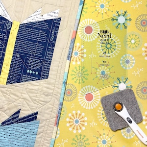 May include: A clear plastic quilting template with the text "Book Nerd Quilt Template" and "A1 Front Cover" and "D1 Back Cover". The template is on a yellow fabric with a white and blue floral pattern.