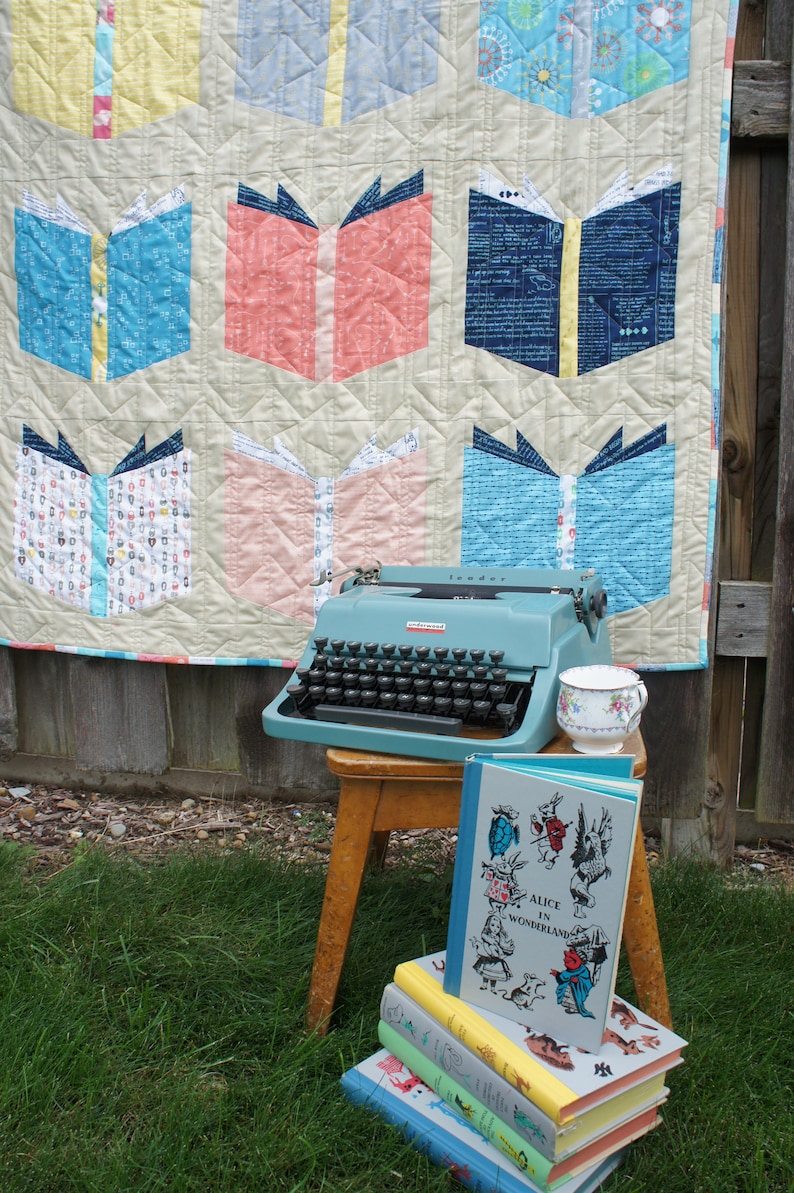 Pu&ograve; includere: Un quilt blu e bianco con un motivo a libro. Una macchina da scrivere vintage blu si trova su uno sgabello di legno con una pila di libri, tra cui "Alice nel Paese delle Meraviglie".
