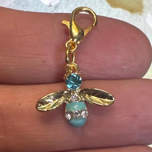 Puede incluir: Un dije de abeja dorado con esmalte turquesa y detalles de cristal transparente. La abeja tiene alas detalladas y un cierre de mosquetón para una fácil fijación. El dije se sostiene entre dos dedos.
