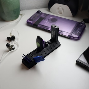 Può includere: Un supporto nero stampato in 3D che contiene vari accessori elettronici, tra cui un'unità USB e un dispositivo di acquisizione HDMI. Una custodia viola per Switch e un controller Xbox bianco e nero sono sullo sfondo. Sono visibili anche auricolari e un telecomando.
