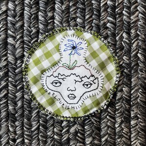 Könnte beinhalten: Ein runder, gestickter Aufnäher mit einem Gesicht und einer Blume, die aus seinem Kopf wächst. Der Aufnäher hat ein grün-weißes Vichy-Muster und die Worte "WIMINA FLOURISHI" sind um das Gesicht herum gestickt. Der Aufnäher befindet sich auf einer strukturierten grau-schwarzen Oberfläche.