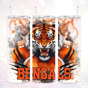 May include: A white tumbler featuring a fierce tiger illustration in orange and black, with the word "BENGALS" in bold orange letters. The design includes a watercolor effect with lightning bolts and a football jersey.
