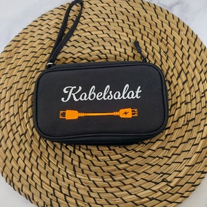Könnte beinhalten: Eine schwarze, rechteckige Aufbewahrungstasche mit Reißverschluss und Handschlaufe. Auf der Vorderseite befindet sich der weiße Schriftzug Kabelsalat über einer orangefarbenen Grafik eines Ladekabels mit Blitzsymbol am Steckerende.