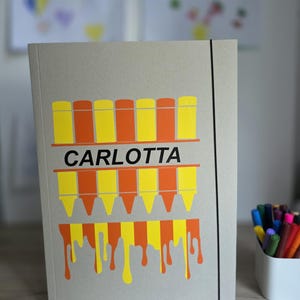 Könnte beinhalten: Eine hellbraune Pappmappe mit schwarzem Gummibandverschluss. Auf der Vorderseite befindet sich eine Grafik aus gelben und orangen Stiften mit dem Namen CARLOTTA in schwarzer Schrift sowie ein tropfender Farbeffekt in Gelb und Orange.