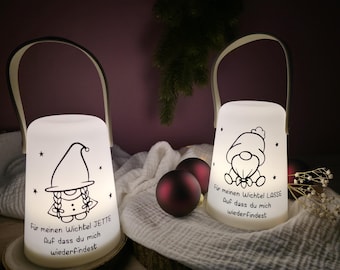 May include: Two white, illuminated lanterns with black handles and gnome illustrations. Each lantern has text in German. The lanterns sit on a wooden surface with Christmas ornaments in the background. The lanterns are cylindrical with a curved handle.