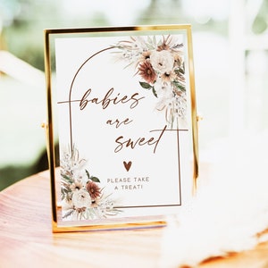 May include: A gold-framed sign with the words "babies are sweet" in elegant script, accented by floral arrangements in neutral tones. The sign also includes the text "PLEASE TAKE A TREAT!" and a heart symbol.