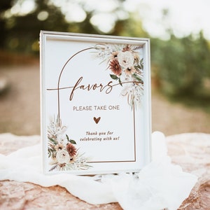 May include: A rectangular white sign with the word "favors" in a cursive font, surrounded by a floral design. The sign also includes the text "PLEASE TAKE ONE" and "Thank you for celebrating with us!"