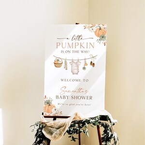 May include: A white rectangular baby shower welcome sign featuring orange pumpkin illustrations, floral accents, and a clothesline graphic with a baby onesie and shoes. The text reads A little PUMPKIN IS ON THE WAY! WELCOME TO Samantha's BABY SHOWER We're so glad you're here!