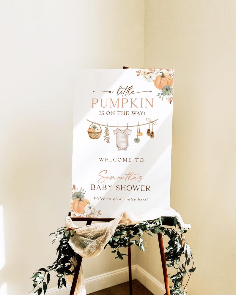 May include: A white rectangular baby shower welcome sign featuring orange pumpkin illustrations, floral accents, and a clothesline graphic with a baby onesie and shoes. The text reads A little PUMPKIN IS ON THE WAY! WELCOME TO Samantha's BABY SHOWER We're so glad you're here!