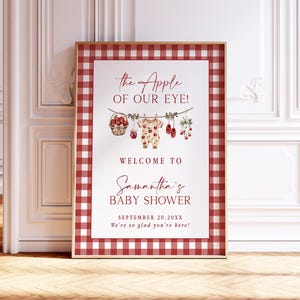 Puede incluir: Un letrero enmarcado para baby shower con un borde de cuadros rojos y blancos. El letrero presenta el texto "the Apple of Our Eye!" y "Welcome to Samantha's Baby Shower." Los elementos decorativos incluyen un body de beb&eacute;, manzanas y zapatos de beb&eacute;.