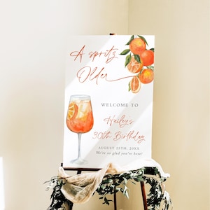 May include: A white sign on an easel with the text "A spritz Older" and "Welcome to Hailey's 30th Birthday". The sign features a watercolor illustration of an orange cocktail and oranges.