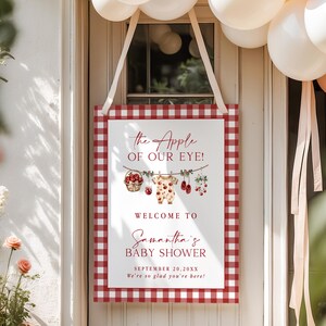 Puede incluir: Un letrero rectangular de baby shower con un borde de cuadros rojos y blancos. El letrero dice "the Apple of Our Eye!" y "Welcome to Samantha's Baby Shower" con la fecha y un mensaje de bienvenida. Decorado con ilustraciones de beb&eacute;.
