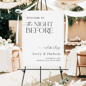 Pu&ograve; includere: Un cartello bianco su un cavalletto nero con il testo "WELCOME TO the NIGHT BEFORE celebrating Avery & Hudson OCTOBER 10, 2026 WE'RE SO GLAD YOU'RE HERE!". Il cartello &egrave; decorato con nastri bianchi e una composizione floreale.