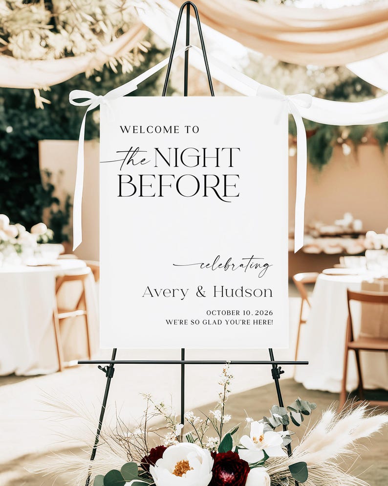 Pu&ograve; includere: Un cartello bianco su un cavalletto nero con il testo "WELCOME TO the NIGHT BEFORE celebrating Avery & Hudson OCTOBER 10, 2026 WE'RE SO GLAD YOU'RE HERE!". Il cartello &egrave; decorato con nastri bianchi e una composizione floreale.