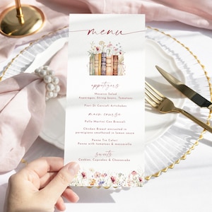 May include: A menu card with a floral and book illustration. The menu lists appetizers, main course, and sweets. The card is held over a white plate with gold cutlery and a pink napkin.