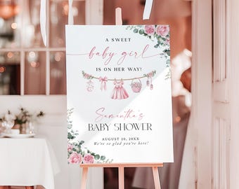 May include: A baby shower sign on a wooden easel. The sign reads "A Sweet Baby Girl Is On Her Way!" in pink script. Floral accents and baby-themed illustrations decorate the sign. White balloons are visible in the background.