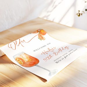 May include: A watercolor-style birthday party sign with an Aperol Spritz illustration. The sign reads "Welcome to Hailey's 30th Birthday" with the date and a welcoming message. The design features orange and red hues.