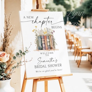 May include: A white sign on a wooden easel with the text "A New Chapter Begins" and "Welcome to Samantha's Bridal Shower". The sign features a watercolor illustration of books and flowers.