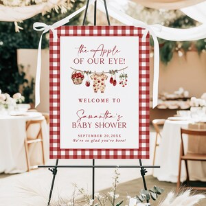 Puede incluir: Un letrero de bienvenida para baby shower con un borde de cuadros rojos y blancos. El letrero dice "the Apple of Our Eye!" y da la bienvenida a los invitados al baby shower de Samantha el 20 de septiembre de 20XX. Elementos decorativos: manzanas y ropa de beb&eacute;.