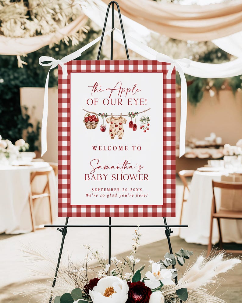 Puede incluir: Un letrero de bienvenida para baby shower con un borde de cuadros rojos y blancos. El letrero dice "the Apple of Our Eye!" y da la bienvenida a los invitados al baby shower de Samantha el 20 de septiembre de 20XX. Elementos decorativos: manzanas y ropa de beb&eacute;.