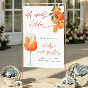 May include: A sign with the text "A Spritz Older" and "Welcome to Hailey's 30th Birthday". The sign features a watercolor illustration of an orange cocktail and oranges. The sign is displayed in a copper-colored frame, with disco balls in the foreground.