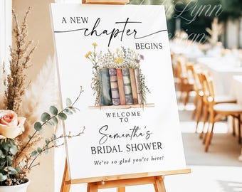May include: A white sign on a wooden easel with the text "A New Chapter Begins" and "Welcome to Samantha's Bridal Shower". The sign features a watercolor illustration of books and flowers. The sign is displayed outdoors.