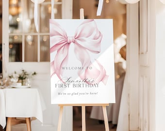 May include: A rectangular welcome sign on a wooden easel featuring a large, soft pink ribbon illustration. The text reads: WELCOME TO Samantha's FIRST BIRTHDAY We're so glad you're here! The sign has a white background and black lettering.