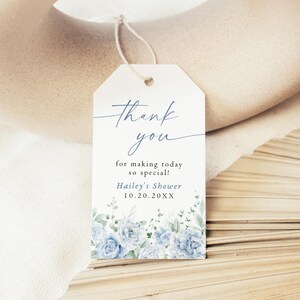 以下が含まれることがあります： 紐付きの長方形の白い紙製ギフトタグ。青い筆記体で「Thank you for making today so special! Hailey's Shower 10.20.20XX」と印刷されており、青い花と緑の葉の水彩画風イラストが下部に描かれています。