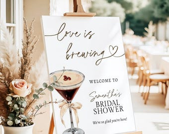May include: A white sign on a wooden easel with the text "love is brewing" and "Welcome to Samantha's Bridal Shower". A watercolor illustration of a cocktail with a bow is featured below the text.
