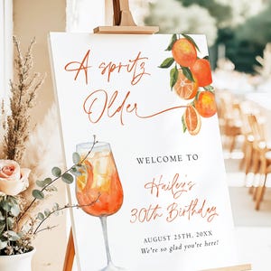 May include: A white sign on a wooden easel with the text "A spritz Older" in orange script. The sign features a watercolor illustration of an orange cocktail and oranges. The sign welcomes guests to Hailey's 30th birthday.