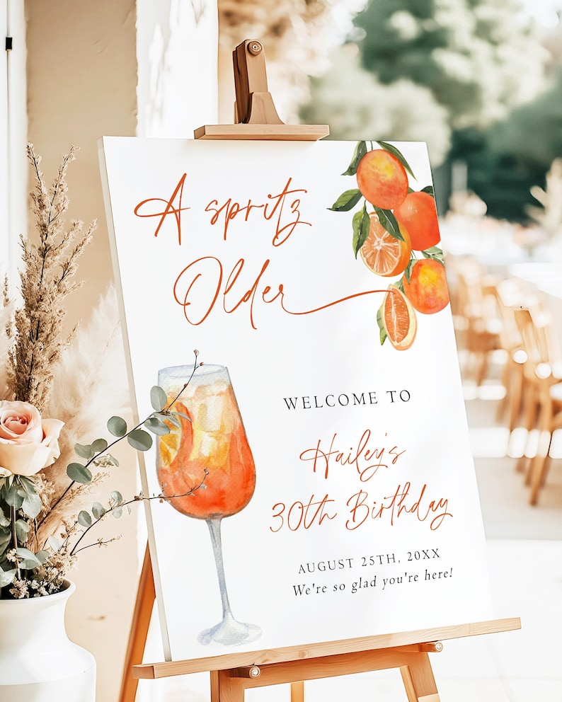 May include: A white sign on a wooden easel with the text "A spritz Older" in orange script. The sign features a watercolor illustration of an orange cocktail and oranges. The sign welcomes guests to Hailey's 30th birthday.