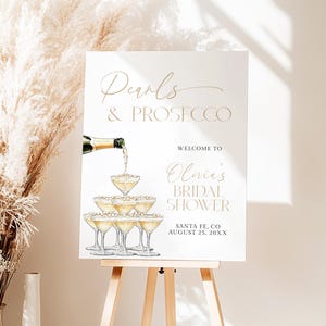 Puede incluir: Un letrero rectangular blanco con el texto Pearls & PROSECCO, WELCOME TO Olivia's BRIDAL SHOWER, SANTA FE, CO AUGUST 25, 20XX. El diseño incluye una ilustración de una botella de champán vertiendo en una torre de copas.