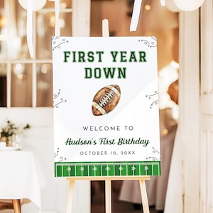 May include: A white sign on a wooden easel with the text "FIRST YEAR DOWN" in green. Below is a football illustration and the text "WELCOME TO Hudson's First Birthday OCTOBER 10, 20XX". The sign has a football field design at the bottom.