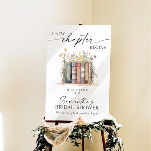 May include: A white sign on an easel with the text "A new chapter begins" and "Welcome to Samantha's Bridal Shower". The sign features a watercolor illustration of books and flowers. Greenery and fabric drape the easel.