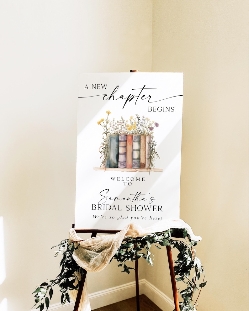 May include: A white sign on an easel with the text "A new chapter begins" and "Welcome to Samantha's Bridal Shower". The sign features a watercolor illustration of books and flowers. Greenery and fabric drape the easel.