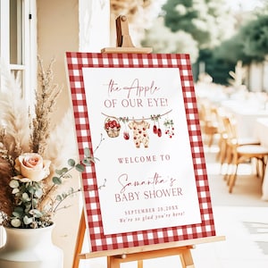 Puede incluir: Un letrero de baby shower con un borde de cuadros rojos y blancos. El letrero dice "The Apple of Our Eye!" y da la bienvenida a los invitados al baby shower de Samantha. Elementos decorativos incluyen manzanas y ropa de beb&eacute;.