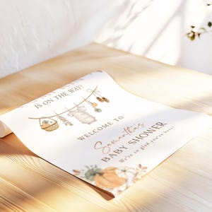 May include: A white rectangular baby shower welcome sign featuring the text IS ON THE WAY, WELCOME TO Samantha's BABY SHOWER, and We're so glad you're here. The design includes illustrations of a clothesline with baby items and pumpkins.