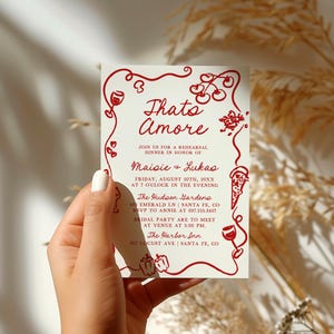 May include: A red and white invitation card with the text "Thats Amore" and details for a rehearsal dinner in honor of Maisie and Lukas. The card features a red illustration of a wine glass, cherries, and a pizza slice.