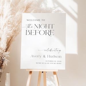 Pu&ograve; includere: Un cartello bianco su un cavalletto di legno con il testo "WELCOME TO The NIGHT BEFORE celebrating Avery & Hudson OCTOBER 10, 2026 WE'RE SO GLAD YOU'RE HERE!" in nero. Lo sfondo &egrave; neutro.