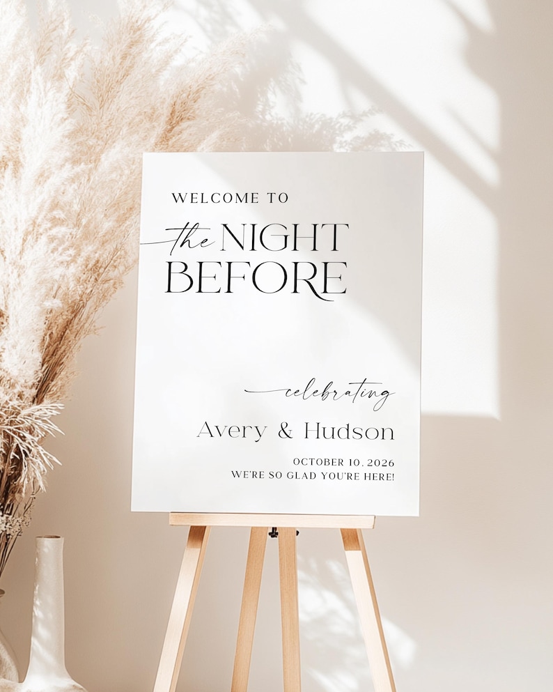 Pu&ograve; includere: Un cartello bianco su un cavalletto di legno con il testo "WELCOME TO The NIGHT BEFORE celebrating Avery & Hudson OCTOBER 10, 2026 WE'RE SO GLAD YOU'RE HERE!" in nero. Lo sfondo &egrave; neutro.