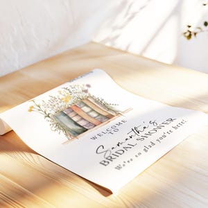 May include: A rolled-up white paper sign with the text "Welcome to Samantha's Bridal Shower". The sign features a watercolor illustration of books and flowers. The sign is on a wooden surface.
