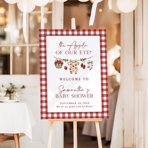 Puede incluir: Un letrero de bienvenida para baby shower sobre un caballete. El letrero tiene un borde a cuadros rojos y blancos y presenta el texto "the Apple of Our Eye!" y "Welcome to Samantha's Baby Shower". Incluye ilustraciones de ropa de beb&eacute; y fruta.