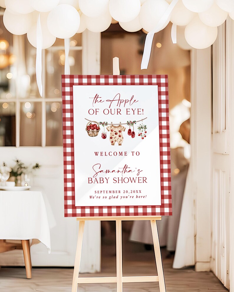 Puede incluir: Un letrero de bienvenida para baby shower sobre un caballete. El letrero tiene un borde a cuadros rojos y blancos y presenta el texto "the Apple of Our Eye!" y "Welcome to Samantha's Baby Shower". Incluye ilustraciones de ropa de beb&eacute; y fruta.