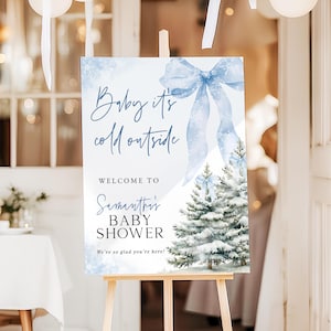 May include: A baby shower sign on an easel. The sign has a winter theme with a blue bow and text that reads "Baby it's cold outside" and "Welcome to Samantha's Baby Shower". White balloons are in the background.