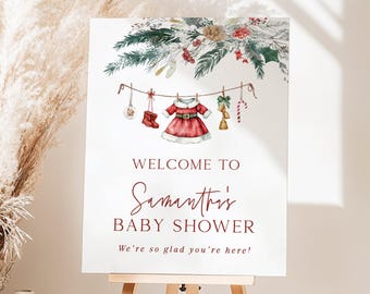 May include: A white rectangular sign featuring a watercolor illustration of pine branches, berries, and a clothesline with a red Santa dress, boots, bells, and a candy cane. Text reads: Welcome to Samantha's Baby Shower. We're so glad you're here!